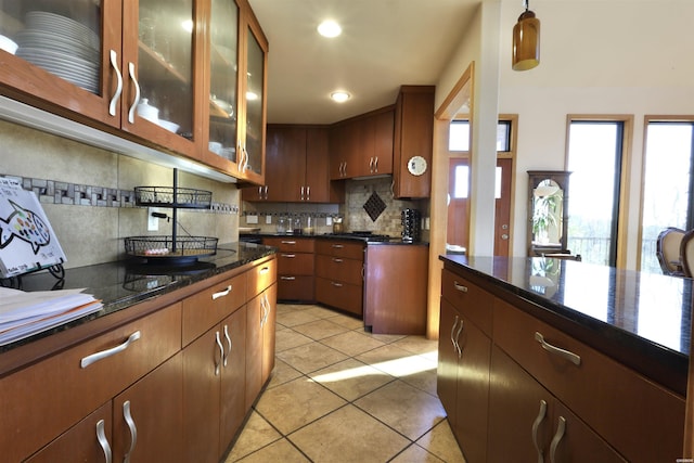 kitchen featuring light tile patterned floors, decorative backsplash, dark stone counters, glass insert cabinets, and pendant lighting