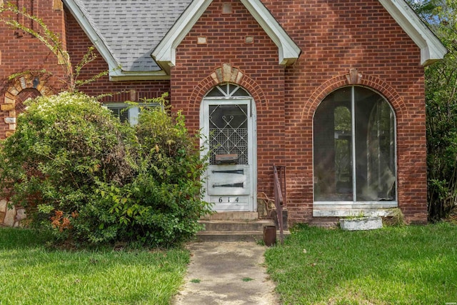 view of exterior entry featuring roof with shingles, a lawn, and brick siding