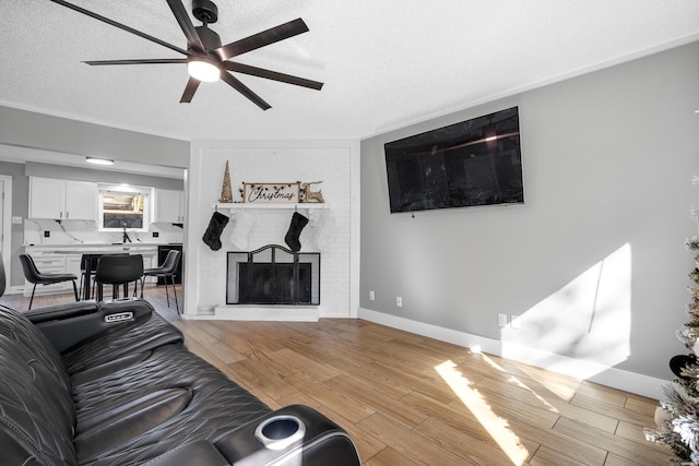 living area with a textured ceiling, ceiling fan, baseboards, a brick fireplace, and light wood finished floors