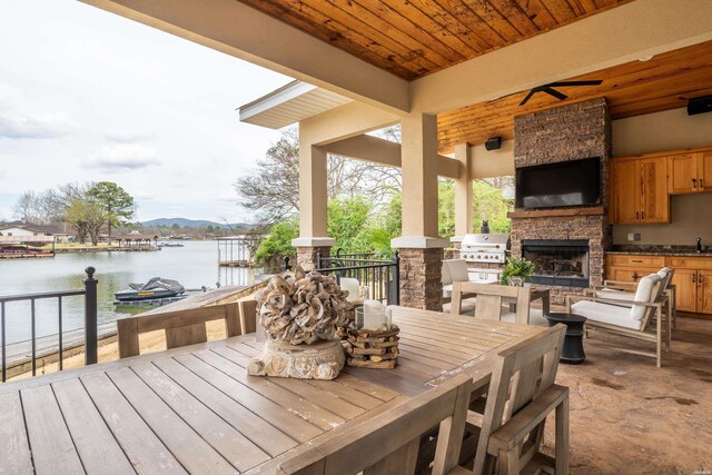 wooden terrace with an outdoor kitchen, an outdoor stone fireplace, a ceiling fan, grilling area, and outdoor dining space