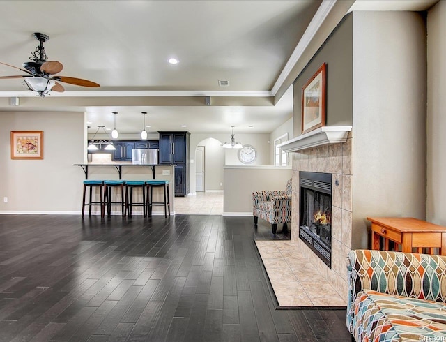 living room with arched walkways, a tiled fireplace, ceiling fan, wood finished floors, and baseboards