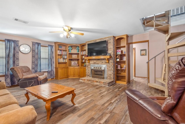 living area featuring visible vents, ceiling fan, stairway, wood finished floors, and a brick fireplace