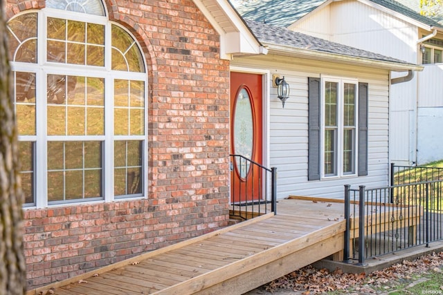 property entrance with brick siding, a shingled roof, a wooden deck, and fence