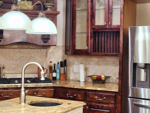 kitchen featuring backsplash, glass insert cabinets, light stone countertops, stainless steel refrigerator with ice dispenser, and a sink