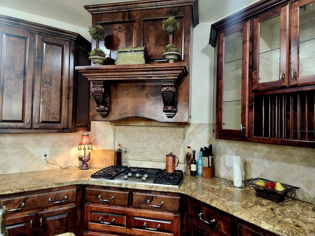 kitchen featuring decorative backsplash, dark brown cabinetry, stainless steel gas stovetop, glass insert cabinets, and light stone countertops