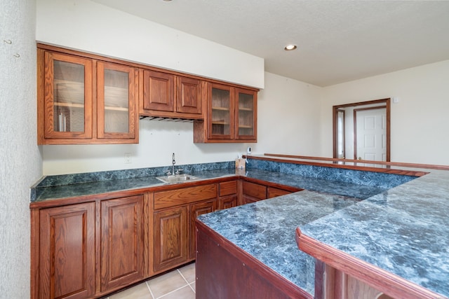 kitchen featuring light tile patterned floors, a sink, brown cabinetry, dark countertops, and glass insert cabinets