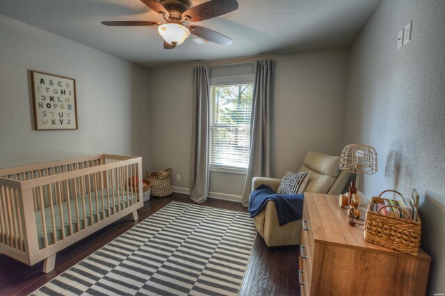 bedroom featuring a crib, dark wood finished floors, a ceiling fan, and baseboards