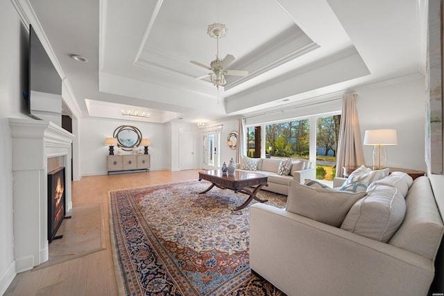 living area with a ceiling fan, a warm lit fireplace, light wood-style floors, crown molding, and a raised ceiling