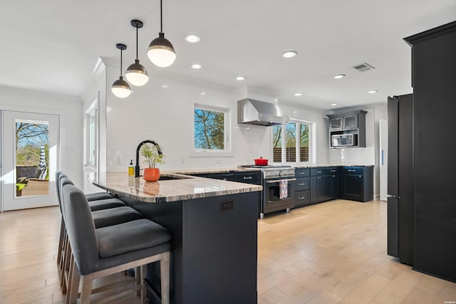 kitchen featuring visible vents, a peninsula, appliances with stainless steel finishes, wall chimney range hood, and light wood-type flooring