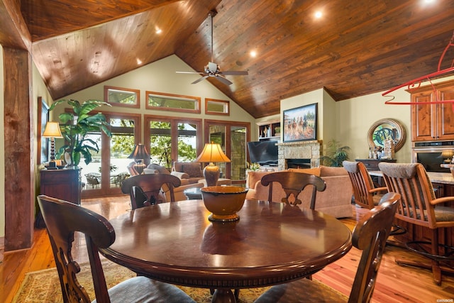 dining space with high vaulted ceiling, a stone fireplace, wood ceiling, a ceiling fan, and light wood-type flooring