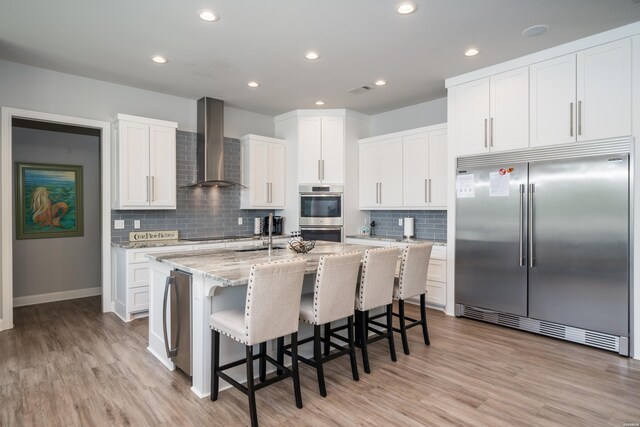 kitchen with appliances with stainless steel finishes, white cabinets, an island with sink, light stone countertops, and wall chimney exhaust hood