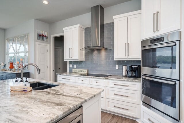 kitchen with black electric stovetop, stainless steel double oven, a sink, white cabinetry, and wall chimney range hood