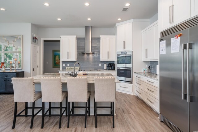 kitchen with a center island with sink, appliances with stainless steel finishes, light stone counters, wall chimney range hood, and white cabinetry