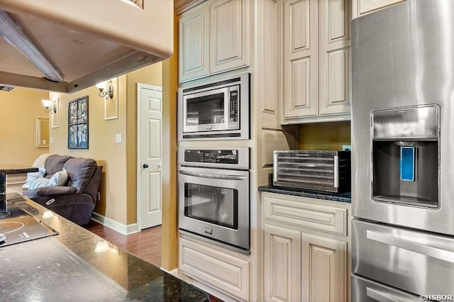 kitchen featuring baseboards, appliances with stainless steel finishes, cream cabinetry, dark stone countertops, and dark wood finished floors