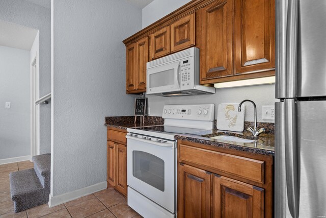 kitchen with light tile patterned floors, brown cabinetry, a sink, dark stone counters, and white appliances