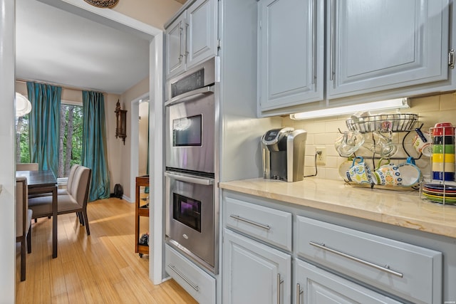 kitchen with stainless steel double oven, tasteful backsplash, light wood-style flooring, and light stone countertops