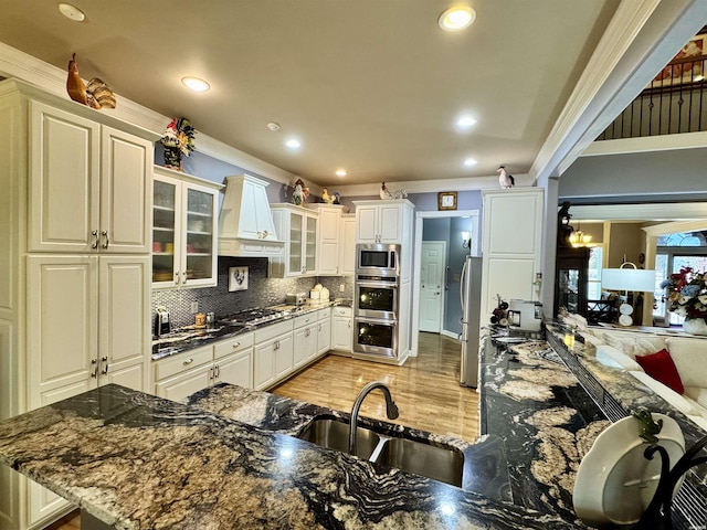 kitchen with glass insert cabinets, dark stone countertops, stainless steel appliances, premium range hood, and a sink