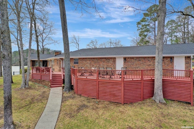 view of front facade with brick siding, roof with shingles, a chimney, a wooden deck, and a front lawn