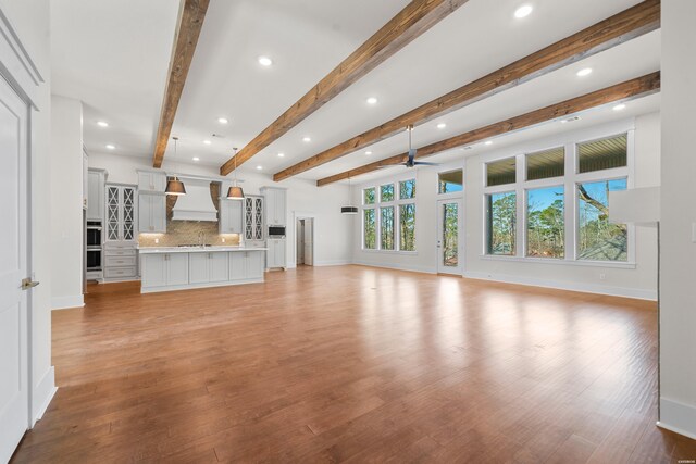 unfurnished living room featuring baseboards, light wood-style flooring, ceiling fan, beamed ceiling, and a sink