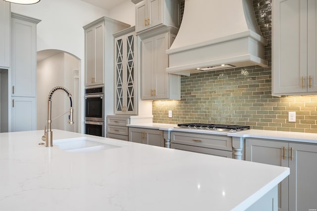 kitchen featuring glass insert cabinets, custom exhaust hood, stainless steel appliances, gray cabinetry, and backsplash
