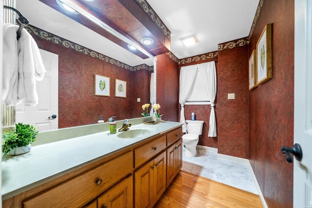 bathroom with toilet, wood finished floors, vanity, baseboards, and wallpapered walls