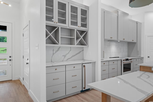 kitchen featuring light stone counters, high quality appliances, light wood-type flooring, decorative backsplash, and glass insert cabinets