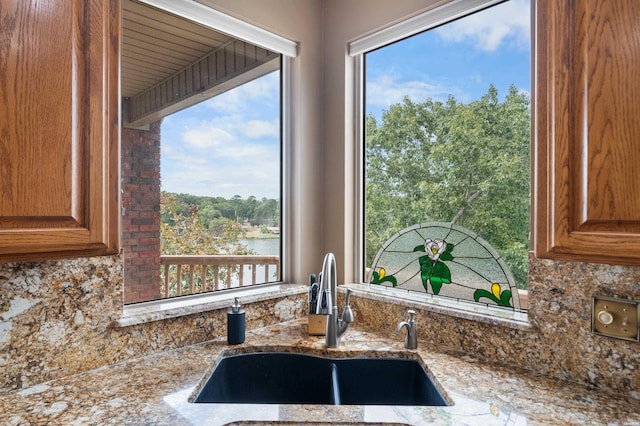 kitchen featuring brown cabinetry, a water view, light stone countertops, a sink, and backsplash