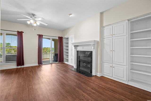 unfurnished living room featuring baseboards, dark wood finished floors, ceiling fan, built in shelves, and a high end fireplace