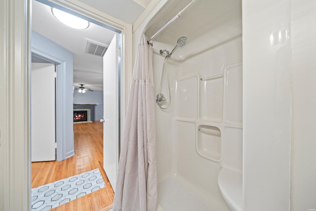 full bathroom with visible vents, ceiling fan, a shower stall, wood finished floors, and a warm lit fireplace