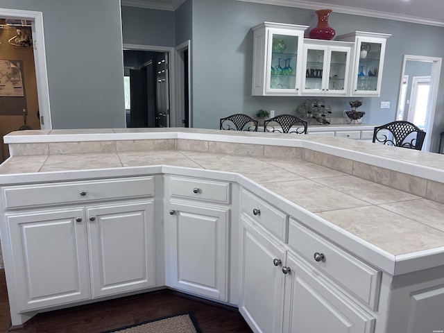 kitchen featuring dark wood finished floors, tile counters, glass insert cabinets, ornamental molding, and white cabinetry