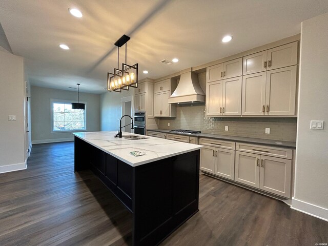 kitchen featuring light stone counters, stainless steel gas cooktop, an island with sink, decorative light fixtures, and custom range hood