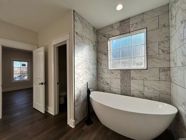 full bath featuring plenty of natural light, a soaking tub, wood finished floors, and toilet