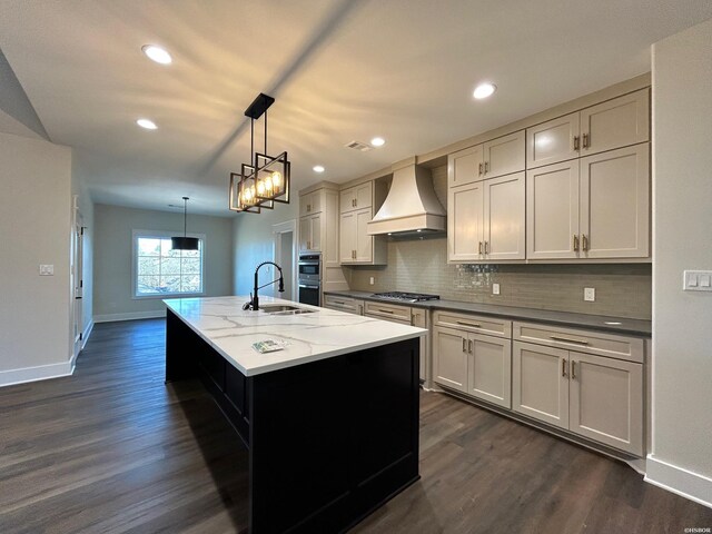 kitchen with stainless steel appliances, premium range hood, a sink, an island with sink, and pendant lighting