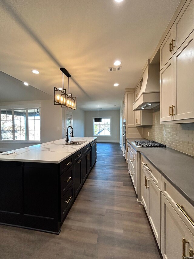 kitchen featuring a kitchen island with sink, premium range hood, white cabinetry, dark cabinetry, and pendant lighting