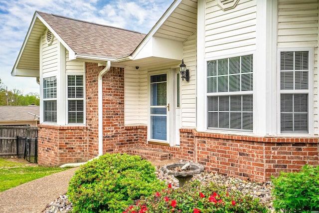 doorway to property featuring brick siding, a shingled roof, and fence