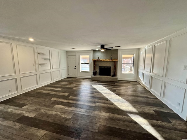 unfurnished living room featuring a brick fireplace, a ceiling fan, a decorative wall, and dark wood-style flooring