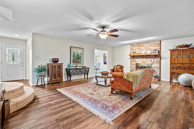 living room featuring dark wood-style flooring, a ceiling fan, a brick fireplace, a textured ceiling, and baseboards