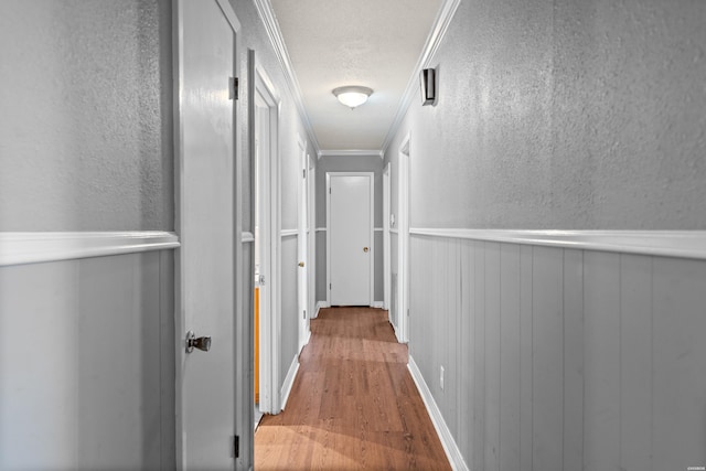 corridor with wood finished floors, a wainscoted wall, ornamental molding, a textured ceiling, and a textured wall