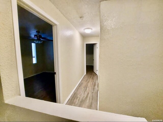 corridor featuring a textured ceiling, a textured wall, wood finished floors, and baseboards