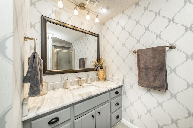 bathroom featuring a textured ceiling, a shower with shower door, vanity, visible vents, and wallpapered walls