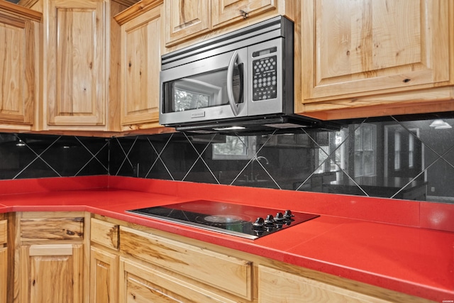 kitchen featuring stainless steel microwave, black electric stovetop, light brown cabinets, backsplash, and dark countertops