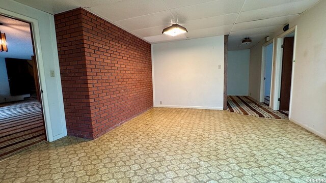 empty room with light floors, baseboards, a drop ceiling, and brick wall