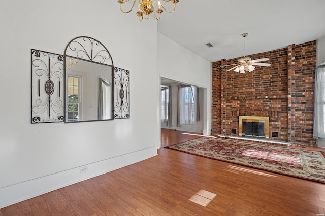 unfurnished living room featuring visible vents, a high ceiling, a brick fireplace, wood finished floors, and ceiling fan with notable chandelier