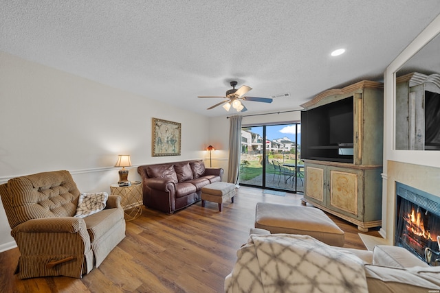 living area featuring a textured ceiling, wood finished floors, visible vents, a ceiling fan, and a lit fireplace