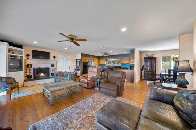 living area featuring a ceiling fan, recessed lighting, a warm lit fireplace, and light wood finished floors