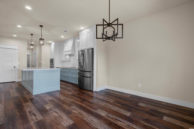 kitchen with decorative light fixtures, custom exhaust hood, light countertops, an island with sink, and stainless steel fridge with ice dispenser