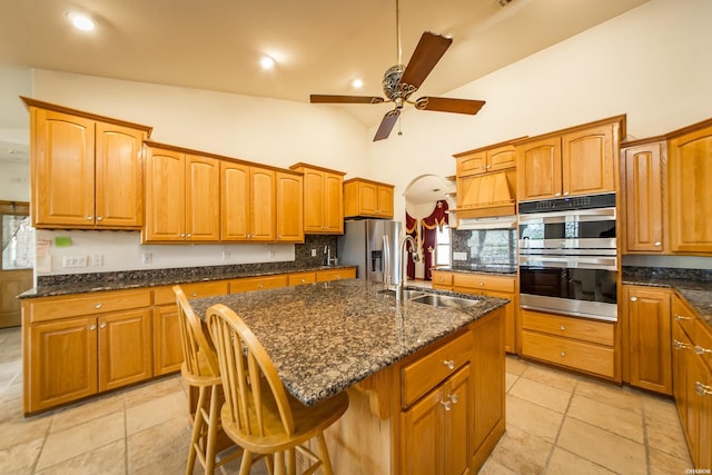 kitchen with brown cabinetry, dark stone counters, an island with sink, a sink, and appliances with stainless steel finishes