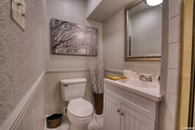 bathroom featuring marble finish floor, a textured wall, toilet, wainscoting, and vanity