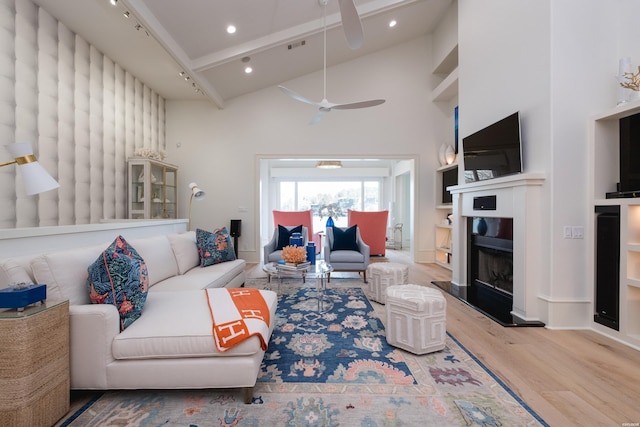 living room with wood finished floors, beamed ceiling, ceiling fan, a fireplace with raised hearth, and high vaulted ceiling