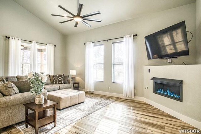 living area featuring ceiling fan, wood finished floors, baseboards, vaulted ceiling, and a glass covered fireplace